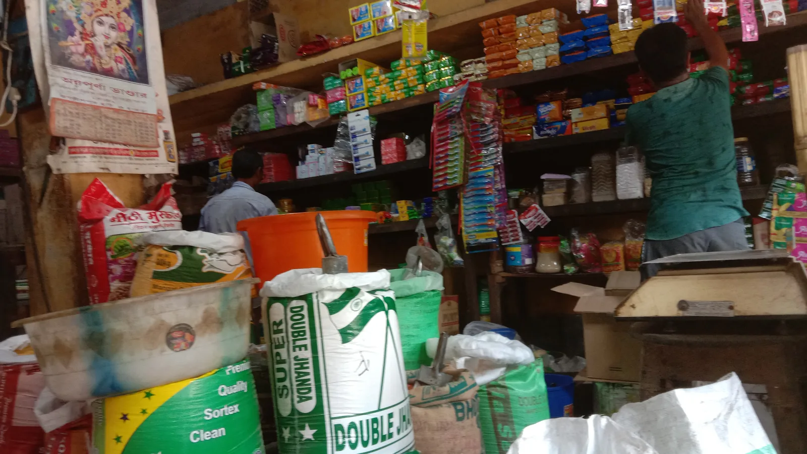 Shopkeepers at the counter of a grocery store in West Bengal, with stock on shelves and a weighing scale for billing