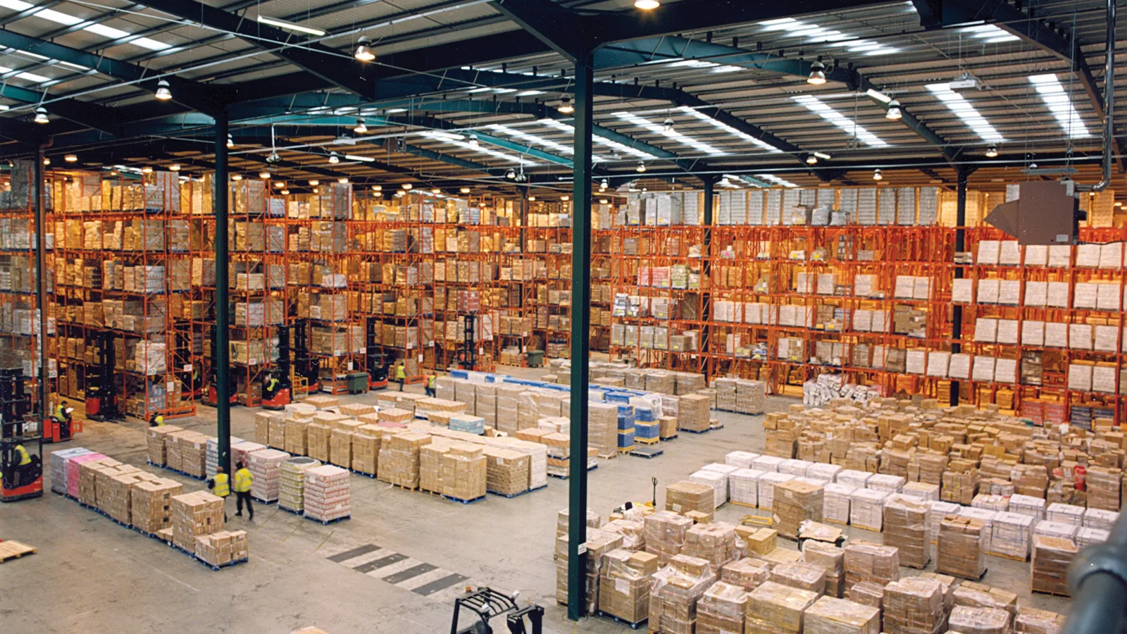 Large warehouse with orange pallet racks, forklifts and stacked cartons of stock