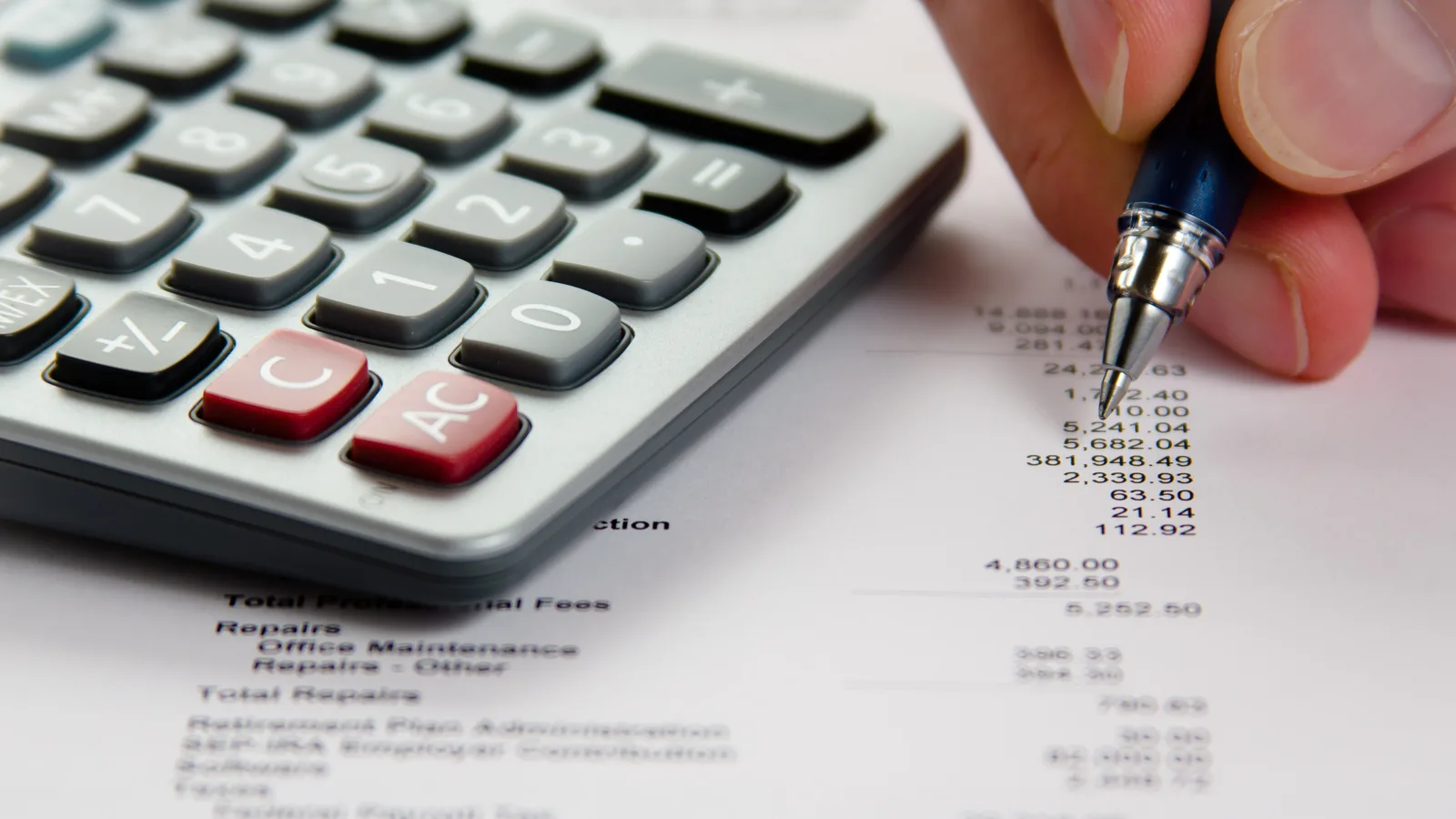 Close-up of a hand with a pen checking figures on a printed financial statement beside a calculator